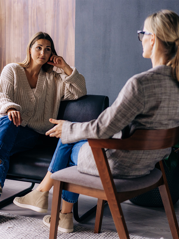 A woman in a cream jumper and jeans sits on a black chair, listening attentively to another woman in a checked blazer and glasses during a conversation in a modern, cosy room.